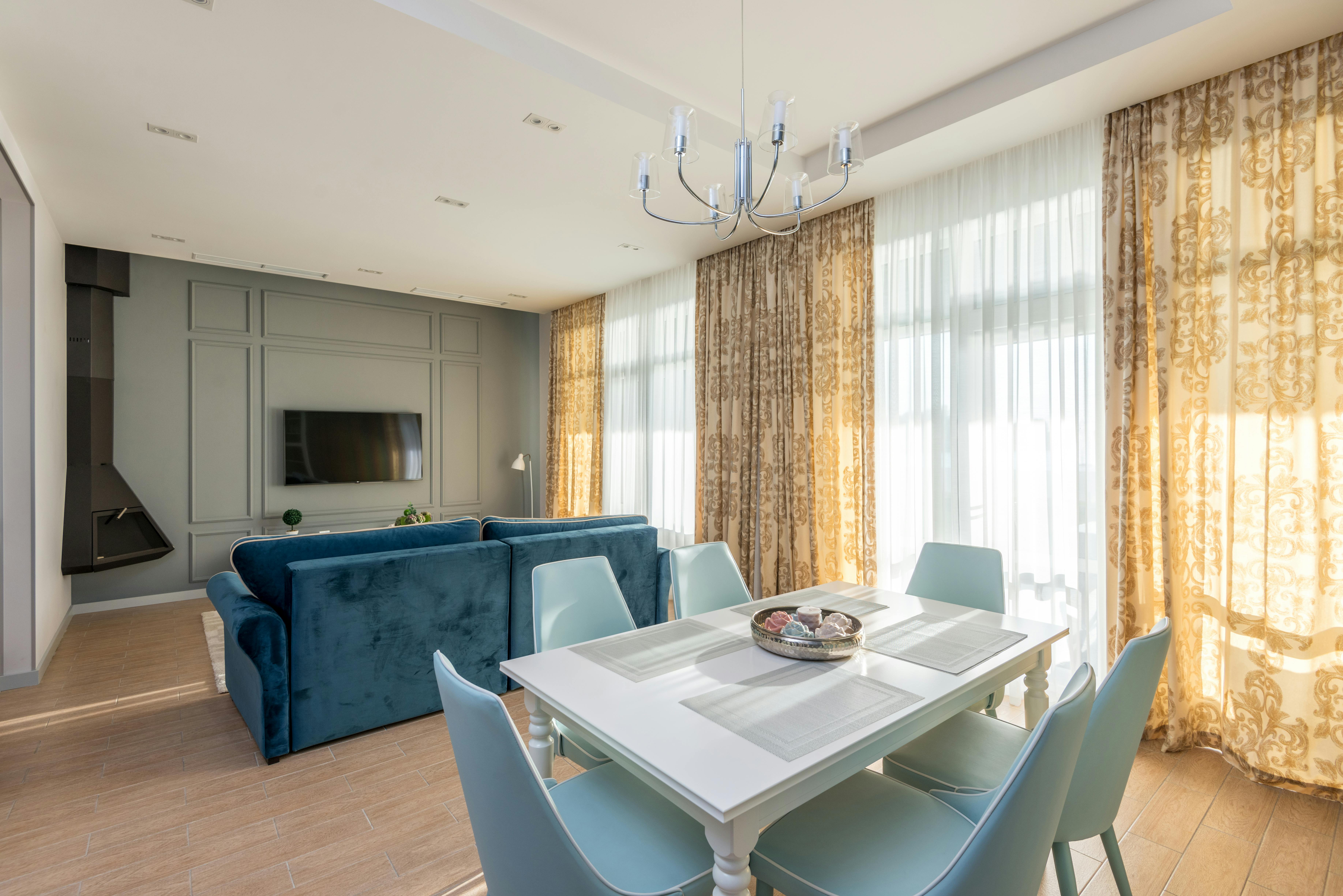 Modern living and dining area with a white table, teal chairs, blue sofa, and TV on a gray wall. Warm light filters through ornate curtains.