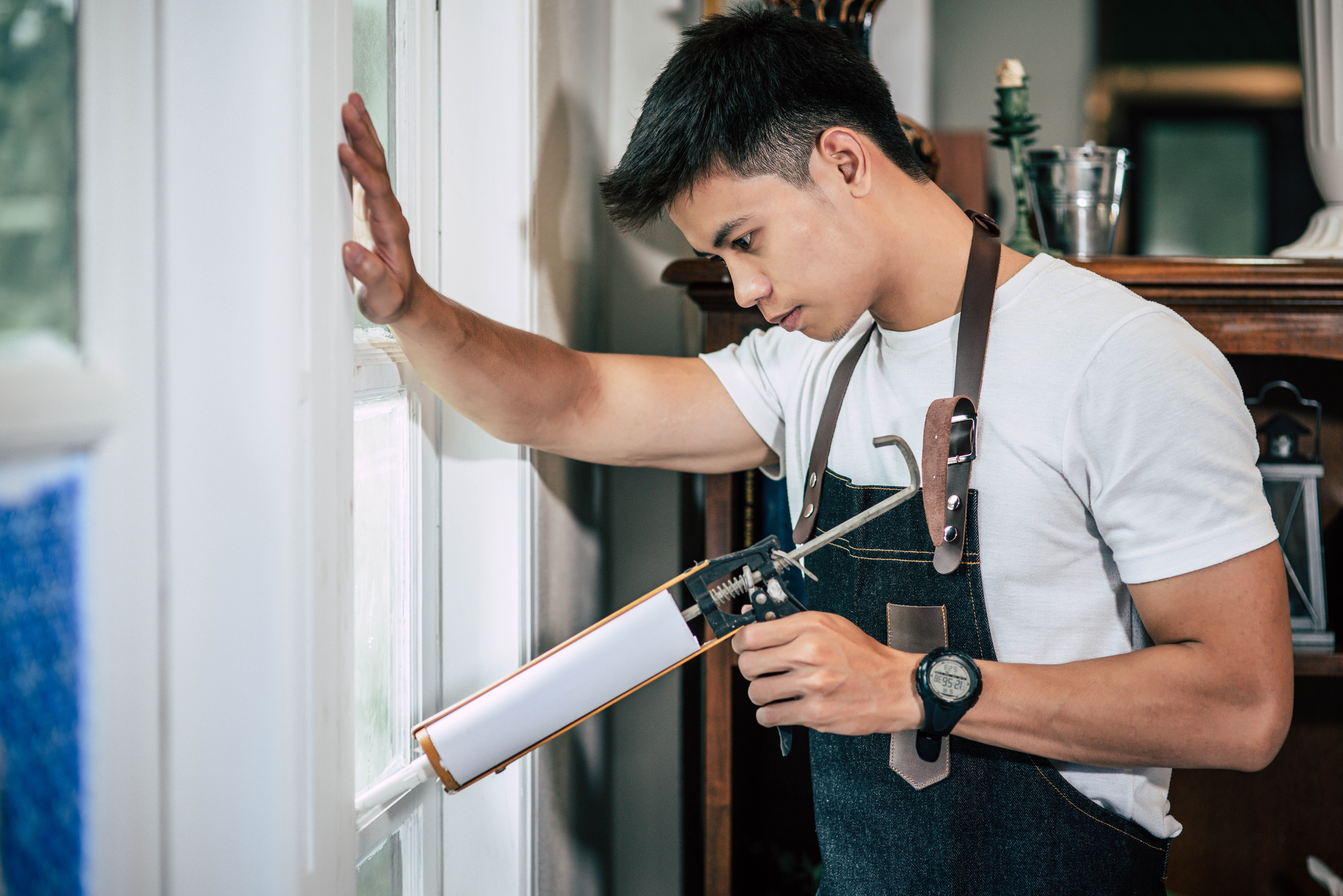 Young man wearing a white t-shirt and apron uses a caulking gun to seal a window frame. He appears focused and concentrated. Indoors, daytime.
